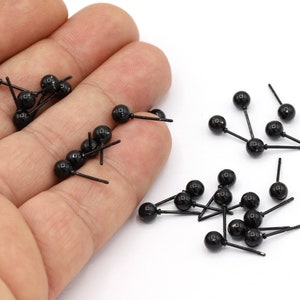 May include: A close-up of a hand holding a pile of black plastic doll eyes with small metal posts. The eyes are scattered on a white surface.