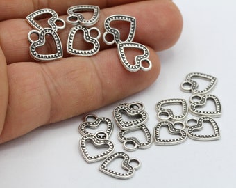 May include: Silver heart-shaped charms with a dotted border and small loop. The charms are scattered on a white surface, some held in a hand. These charms are suitable for jewellery making and crafting.