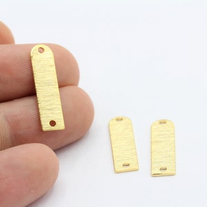 May include: Gold-colored rectangular metal tags with a textured surface and rounded top. Each tag has a hole at the top and bottom. One tag is held by a hand, with two others lying on a white surface.