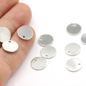 Puede incluir: Una colección de diez dijes redondos de metal plateado con un pequeño agujero en la parte superior. Los dijes son lisos y tienen una superficie ligeramente texturizada.