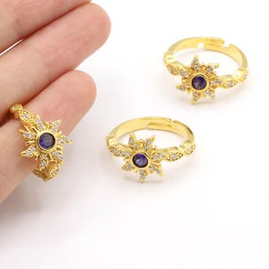 Op de afbeelding: Verstelbare goudkleurige ringen met een zonnestraalontwerp. Elke ring heeft een centrale donkerblauwe steen omringd door kleine, heldere kristallen. De band is gedetailleerd met kleine, getextureerde patronen. De ringen worden op een witte achtergrond weergegeven.
