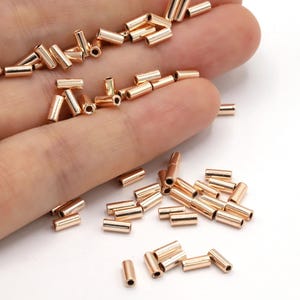 May include: A selection of small, cylindrical, rose gold-coloured metal beads. The beads are scattered across a hand, with some resting on a white surface. These components are often used in jewellery making and crafting.