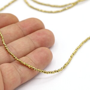 May include: A gold-toned necklace made of small, faceted beads. The necklace is displayed on a hand, showing the length and detail of the beads.