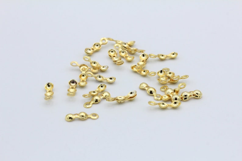 May include: A group of gold-colored metal bead caps, each with a small loop on one end and two rounded ends. The caps are scattered on a white surface.