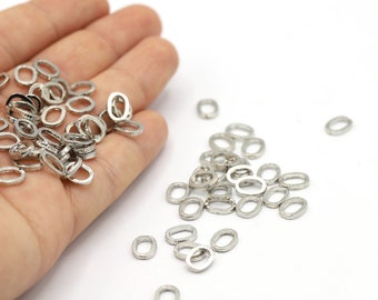 May include: A collection of small, silver-colored oval-shaped metal rings. Some rings are held in a hand, while others are scattered on a white surface. The rings have a textured, hammered appearance, suggesting a handmade or vintage style. Ideal for jewelry making.