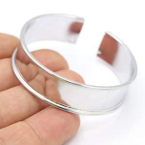Peut inclure: Un bracelet jonc en métal argenté avec une bande large et plate. Le bracelet est ouvert d'un côté et a une finition lisse et polie.