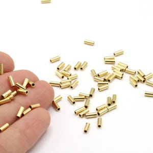 Può includere: Una collezione di piccole perline cilindriche in metallo color oro sparse su una superficie bianca. Alcune perline sono tenute in mano, mentre altre sono sparse. Queste perline sono probabilmente utilizzate per la creazione di gioielli o altri progetti artigianali.