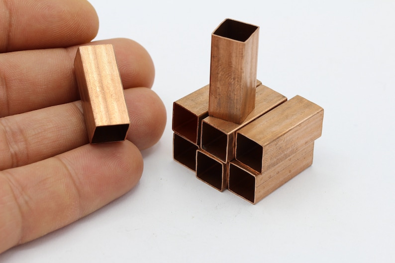 May include: A group of small, copper-colored, square-shaped tubes stacked together. One tube is held in a hand for size comparison.