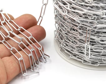 May include: A spool of silver-colored chain with rectangular links. The chain is lying on a white surface and a hand is holding a section of the chain.