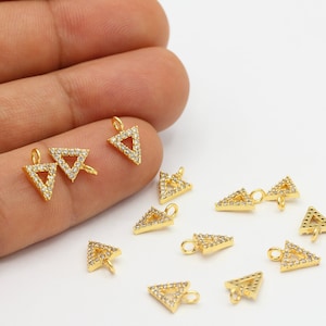 Peut inclure: Charm triangulaire doré orné de strass transparents. Les breloques ont une forme triangulaire et une boucle pour l'attache. Plusieurs breloques sont disposées sur une surface blanche.