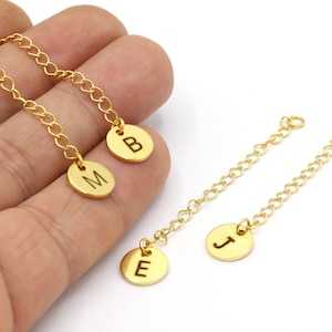 May include: Two gold chains with initial charms. The chains are on a white background. The charms are round and have the letters "E", "J", "M", and "B" engraved on them.