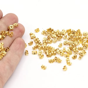 Pode incluir: Uma grande quantidade de fechos de brincos dourados. Os fechos são feitos de metal e têm um pequeno laço de cada lado. Eles estão espalhados sobre uma superfície branca.