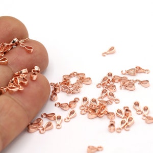 May include: A large quantity of rose gold-colored metal pendant bails. The bails are small and have a teardrop shape. They are scattered on a white surface.