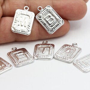 Puede incluir: Pendientes de plata con forma de encanto con las letras M, B, T, A, E e I. Cada encanto es rectangular con un borde elevado y una letra en el centro.