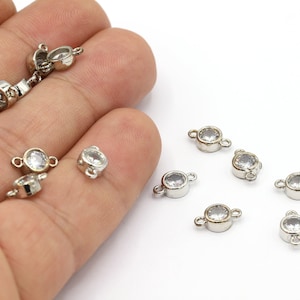 May include: Silver-toned jewelry findings with clear, round, faceted stones. The findings have small loops on either side for attaching to other components. The image shows several of these findings, some held in a hand, against a white background.