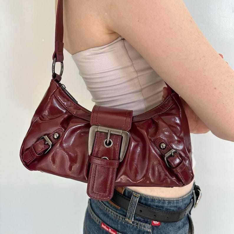 Pu&ograve; includere: Una borsa a spalla bordeaux lucida con chiusura a fibbia argentata. La borsa presenta una forma curva, un cinturino corto e dettagli hardware argentati. La borsa &egrave; tenuta da una persona.