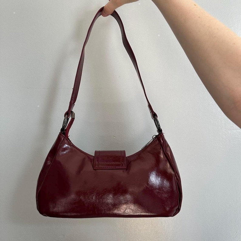 Pu&ograve; includere: Una borsa a spalla bordeaux con finitura lucida. La borsa presenta una forma curva, una singola tracolla e una chiusura rettangolare. La tracolla &egrave; attaccata alla borsa con anelli metallici color argento.