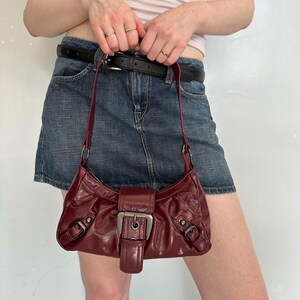 Pu&ograve; includere: Una borsa a tracolla in pelle bordeaux con chiusura a fibbia argentata. La borsa ha una forma curva e un cinturino corto. La borsa &egrave; indossata a spalla. La borsa &egrave; realizzata in pelle lucida.