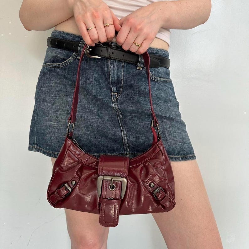 Pu&ograve; includere: Una borsa a tracolla in pelle bordeaux con chiusura a fibbia argentata. La borsa ha una forma curva e un cinturino corto. La borsa &egrave; indossata a spalla. La borsa &egrave; realizzata in pelle lucida.
