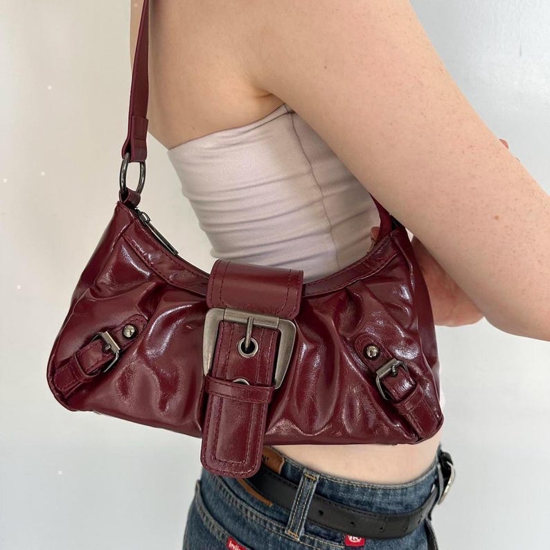 Pu&ograve; includere: Una borsa a spalla in vernice bordeaux con chiusura a fibbia color argento. La borsa ha una forma curva e una singola tracolla. La borsa &egrave; larga circa 25 cm.