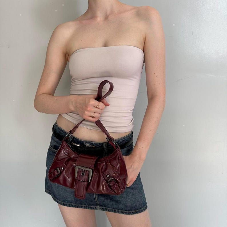 Pu&ograve; includere: Borsa a spalla in pelle bordeaux con chiusura a fibbia argentata. La borsa ha una forma curva e un manico corto. La persona indossa un top a tubo rosa chiaro e una minigonna di jeans.