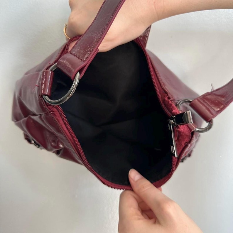 Pu&ograve; includere: Una borsa a spalla bordeaux con finitura lucida. La borsa presenta uno scomparto principale con cerniera, anelli in metallo color argento e una tracolla abbinata. L'interno &egrave; nero. La borsa &egrave; tenuta aperta, rivelando l'interno.