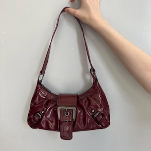 Pu&ograve; includere: Borsa a spalla bordeaux con finitura lucida. La borsa presenta una chiusura con fibbia color argento e due fibbie pi&ugrave; piccole sui lati. Il cinturino &egrave; in tinta bordeaux.