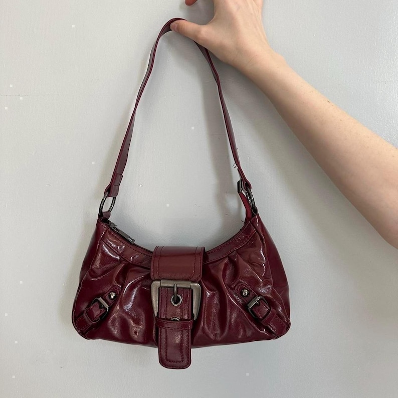 Pu&ograve; includere: Borsa a spalla bordeaux con finitura lucida. La borsa presenta una chiusura con fibbia color argento e due fibbie pi&ugrave; piccole sui lati. Il cinturino &egrave; in tinta bordeaux.