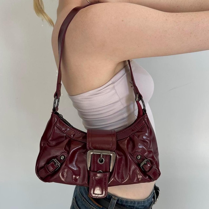 Pu&ograve; includere: Una borsa a spalla bordeaux con finitura lucida. La borsa presenta una grande fibbia argentata e fibbie pi&ugrave; piccole sui lati. Anche il cinturino &egrave; bordeaux. La borsa ha una forma a mezzaluna.