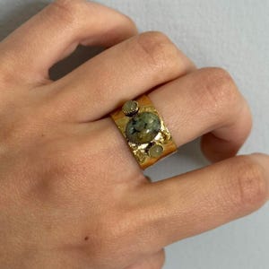May include: A gold-colored ring with a textured band. The ring features an oval-shaped, green-speckled stone in the center, flanked by two smaller, round, light-colored stones. The ring is worn on a finger.