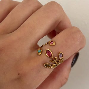 May include: A gold-toned ring with a leaf design, adorned with colorful gemstones. The ring features a branch-like structure with a large, oval ruby-colored stone and smaller, round gemstones in shades of pink, blue, and green. The ring is on a finger.