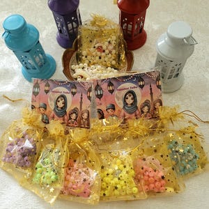 May include: Assortment of colorful beaded prayer strings in small organza bags with gold stars. The bags are arranged in a semi-circle around two cards with the text "Nasamat Taiba" in Arabic script. The cards are surrounded by five miniature lanterns in various colors, including blue, purple, red, and white.