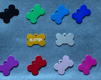 May include: A collection of colorful dog bone-shaped pet tags. The tags are in various colors, including black, green, blue, purple, red, and silver. One gold tag has the name "BUSTER" engraved on it. Each tag has a small hole for attachment.