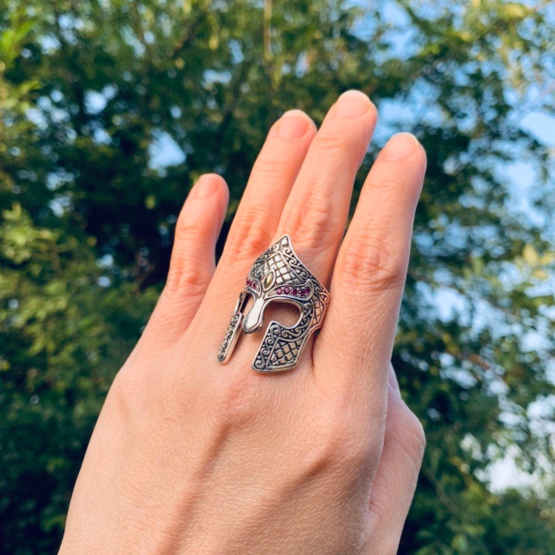 Puede incluir: Un anillo de plata con un dise&ntilde;o detallado de un casco espartano. El casco est&aacute; decorado con peque&ntilde;as piedras preciosas rojas.