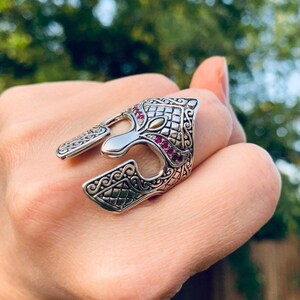 Puede incluir: Un anillo de plata con un dise&ntilde;o detallado de un casco de guerrero. El casco est&aacute; adornado con piedras preciosas rosas y un acento dorado.