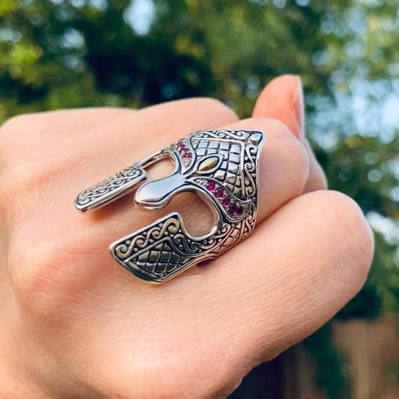 Puede incluir: Un anillo de plata con un dise&ntilde;o detallado de un casco de guerrero. El casco est&aacute; adornado con piedras preciosas rosas y un acento dorado.