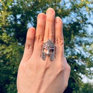 Puede incluir: Un anillo de plata con un dise&ntilde;o detallado de un casco de guerrero. El casco est&aacute; decorado con peque&ntilde;as piedras preciosas rojas.