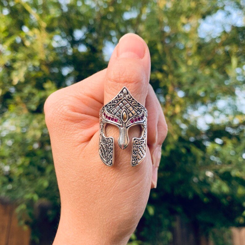 Puede incluir: Un anillo de plata con un dise&ntilde;o detallado de un casco de guerrero. El casco est&aacute; adornado con piedras preciosas rojas y un acento dorado.