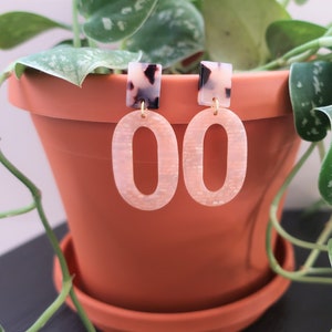 May include: A pair of pink oval earrings with a white and black tortoise shell accent. The earrings have a gold-tone finish.