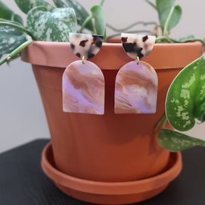 Puede incluir: Pendientes colgantes con diseño veteado. Los pendientes presentan una parte superior semicircular en patrón carey, conectada a una forma de arco más grande en tonos lavanda, marrón y crema. Cuelgan de postes dorados.