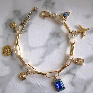 May include: A gold charm bracelet with a variety of charms, including a blue eye, a small airplane, a globe, a camera, and a blue gemstone. The bracelet is made of gold-toned metal and has a lobster clasp.