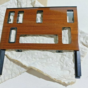 Może przedstawiać: Prostokątny panel z drewnianym wzorem, z wieloma prostokątnymi wycięciami i czarnymi obramowaniami. Panel ma dwa czarne metalowe wsporniki u dołu. Panel spoczywa na pogniecionej, kremowej powierzchni.