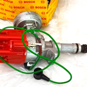 Puede incluir: Un distribuidor automotriz nuevo con tapa roja, cuerpo plateado y conector negro. Un cable verde rodea el distribuidor. La caja en el fondo tiene la marca "BOSCH" impresa.