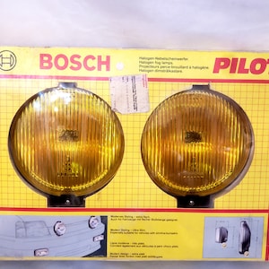May include: A pair of round yellow fog lights in a yellow and black box. The box has the Bosch logo and the text "Halogen Nebelscheinwerfer. Halogen fog lamps. Projecteurs perce-brouillard à halogène Halogen dimstrålkastare. PILOT" printed on it.