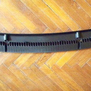May include: Black plastic car part with a curved shape and a series of small holes. The part has two small black tabs on the sides.