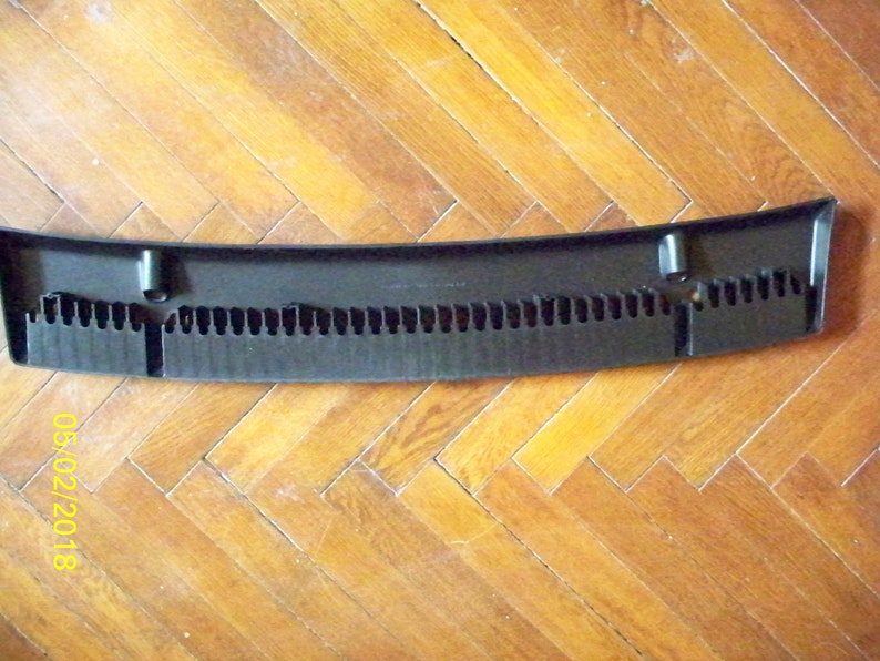 May include: Black plastic car part with a curved shape and a series of small holes. The part has two small black tabs on the sides.