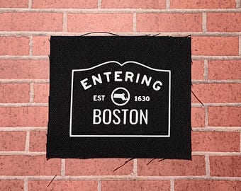 May include: Black fabric patch with white text that reads "Entering Est. 1630 Boston" with a white outline of the state of Massachusetts inside a circle.