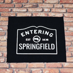 May include: Black fabric patch with white text that reads "Entering Springfield Est. 1636" with a circle containing the outline of the state of Massachusetts.