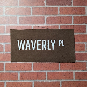 Könnte beinhalten: Ein braunes Canvas-Schild mit weißem Text, auf dem "WAVERLY PL" steht.