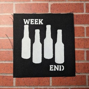 Op de afbeelding: Zwarte stoffen patch met witte tekst en afbeelding. De tekst luidt "WEEK END" met vier witte silhouetten van bierflessen tussen de woorden.